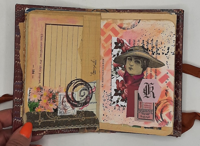 May include: Open vintage journal with aged paper pages. Features a collage of stamps, floral accents, and a portrait of a woman in a hat. Includes handwritten text and decorative elements.