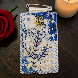 May include: A handmade fabric pin with a patchwork design. The pin features blue floral patterns, white lace, and embroidered details. A yellow flower and a blue tree design are stitched on the fabric. A safety pin is attached to the top.