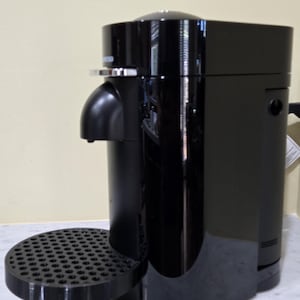 May include: A black espresso machine with a glossy finish. The machine features a curved spout, a round drip tray, and a cylindrical body. The design is modern and compact, suitable for home use.