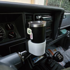 May include: A black and white cup holder with a black travel mug inside. The cup holder is mounted to the dashboard of a car. The travel mug has a tea bag hanging from the lid.