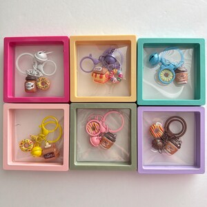 May include: Six square display frames in pink, yellow, teal, light pink, sage green, and lavender, each containing a keychain with miniature food charms including donuts, coffee cups, and small bells, arranged in two rows of three.
