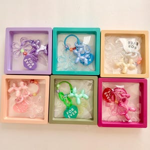 May include: Six square display boxes in purple, teal, beige, peach, olive green, and magenta, each containing a keychain. The keychains feature a balloon animal figure, a clear flower-shaped charm, and a heart-shaped tag with the text FOR EVER.