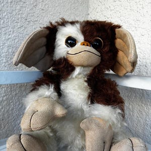 May include: A plush toy character with brown and white fur, large dark eyes, and oversized tan ears. The creature has a tan face, hands, and feet with stitched details. The right foot features the text LTD. printed in black ink on the surface.