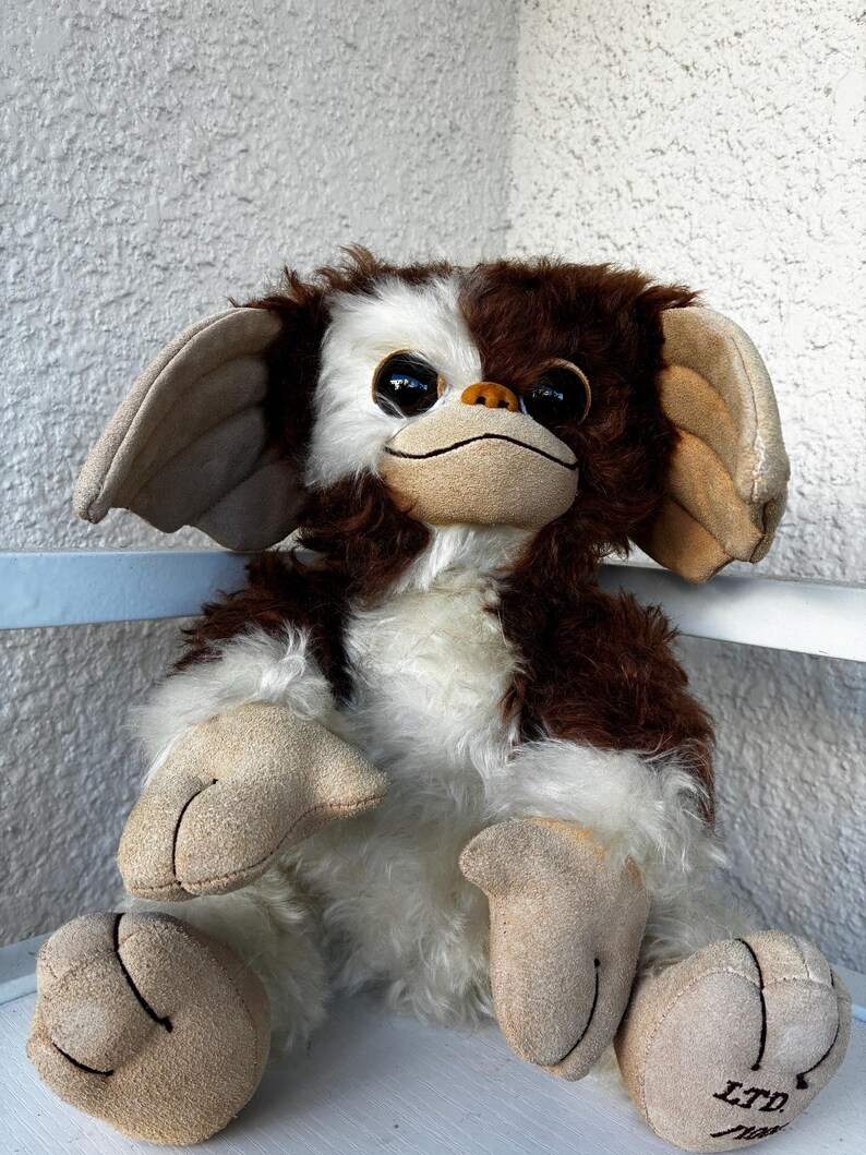 May include: A plush toy character with brown and white fur, large dark eyes, and oversized tan ears. The creature has a tan face, hands, and feet with stitched details. The right foot features the text LTD. printed in black ink on the surface.