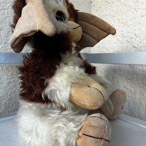 May include: A plush toy featuring a creature with brown and white fur, large pointed ears, and tan suede-like paws. The figure has a rounded face with dark eyes and a small nose, sitting in an upright position with its limbs tucked forward.