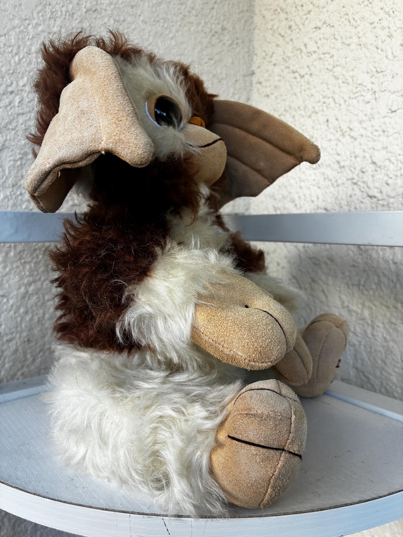 May include: A plush toy featuring a creature with brown and white fur, large pointed ears, and tan suede-like paws. The figure has a rounded face with dark eyes and a small nose, sitting in an upright position with its limbs tucked forward.