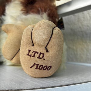 May include: A plush toy with long, shaggy brown and cream-colored fur. The focus is on the tan, suede-like foot pad, which features the dark brown embroidered text 'LTD.' above '/1000', indicating a limited edition production run of this item.