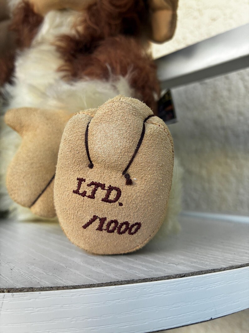 May include: A plush toy with long, shaggy brown and cream-colored fur. The focus is on the tan, suede-like foot pad, which features the dark brown embroidered text 'LTD.' above '/1000', indicating a limited edition production run of this item.