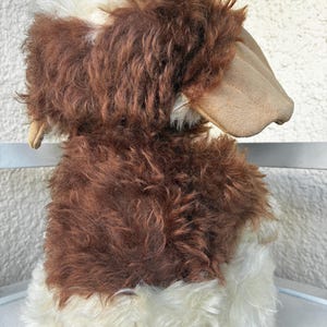 May include: A plush stuffed animal featuring long, shaggy fur in shades of dark brown and cream. The toy has large, smooth, tan-colored ears that hang down on the sides. The texture of the fur is thick and curly, covering the body and head.