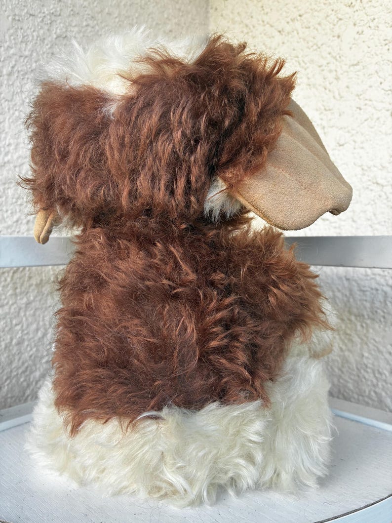 May include: A plush stuffed animal featuring long, shaggy fur in shades of dark brown and cream. The toy has large, smooth, tan-colored ears that hang down on the sides. The texture of the fur is thick and curly, covering the body and head.
