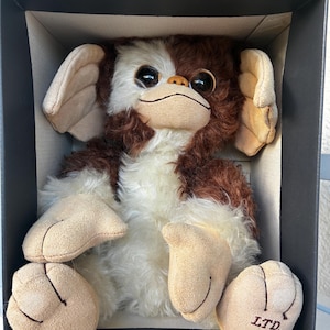 May include: A plush creature with brown and white fur, large dark eyes, and tan suede-like ears, hands, and feet. The creature is seated inside a black box. The right foot features the text LTD. /1000 embroidered in dark brown thread.