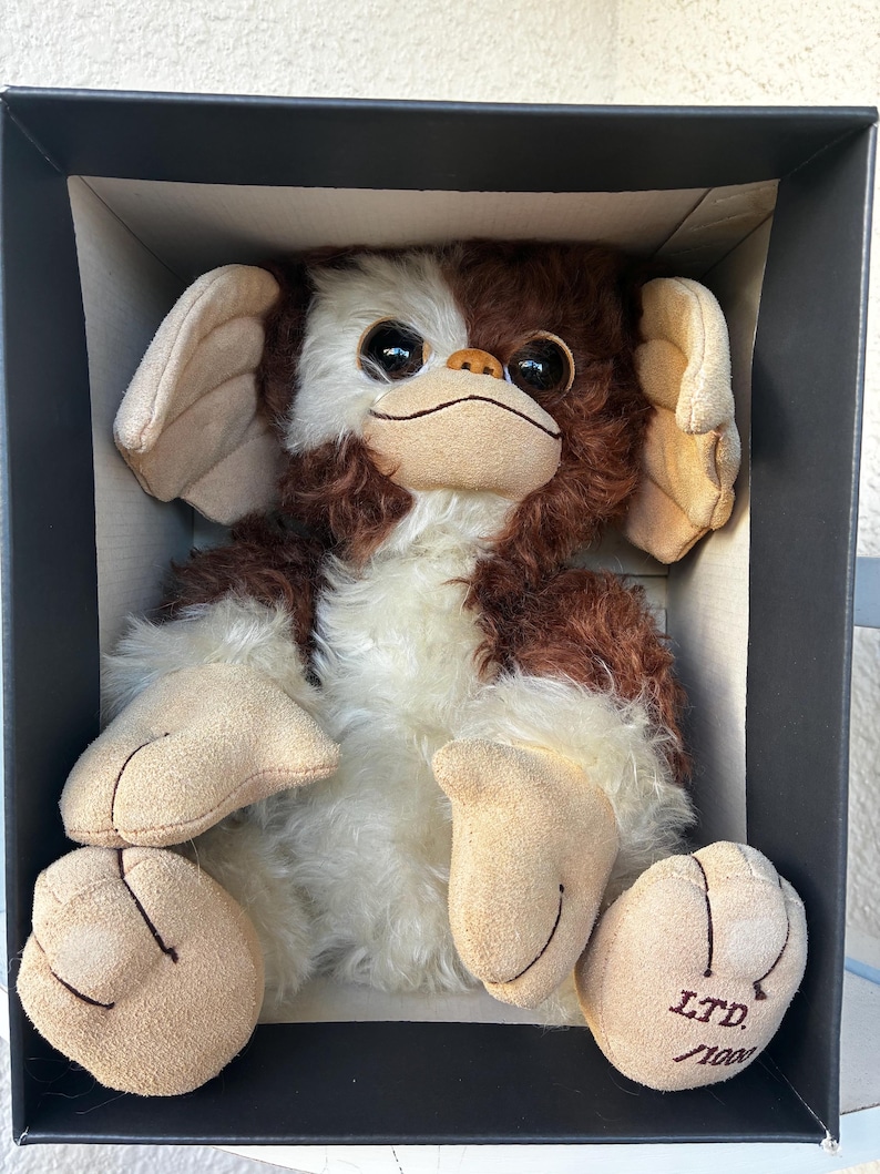 May include: A plush creature with brown and white fur, large dark eyes, and tan suede-like ears, hands, and feet. The creature is seated inside a black box. The right foot features the text LTD. /1000 embroidered in dark brown thread.
