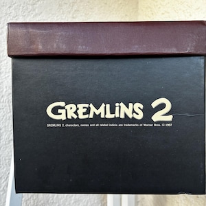 May include: A rectangular black cardboard box with a dark brown lid. The front features the text GREMLINS 2 in a cream-colored font, followed by the smaller printed text: GREMLINS 2, characters, names and all related indicia are trademarks of Warner Bros. &copy; 1997.