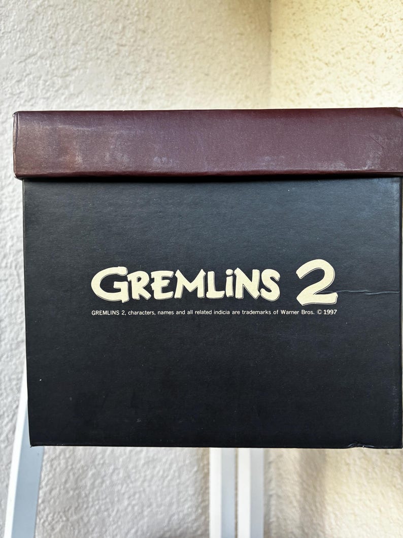 May include: A rectangular black cardboard box with a dark brown lid. The front features the text GREMLINS 2 in a cream-colored font, followed by the smaller printed text: GREMLINS 2, characters, names and all related indicia are trademarks of Warner Bros. &copy; 1997.