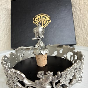 Puede incluir: Un tapón de botella plateado con Tweety Bird, sobre un corcho, con una base floral decorativa. El tapón está colocado en una bandeja circular plateada y ornamentada con un interior negro. Una caja negra con el logo de WB está en el fondo.