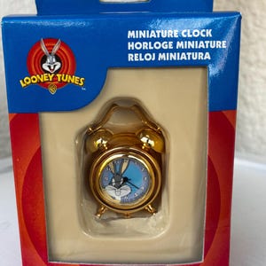 May include: A gold-toned miniature alarm clock featuring a Bugs Bunny graphic on the clock face, housed in a blue and red Looney Tunes branded box with the text Miniature Clock, Horloge Miniature, and Reloj Miniatura printed on the front.