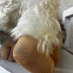 May include: A plush toy featuring long, shaggy white faux fur covering the upper portion of the limbs, transitioning into smooth, tan-colored suede-like fabric at the hooves. The hooves are split, showing a distinct cleft design at the base.