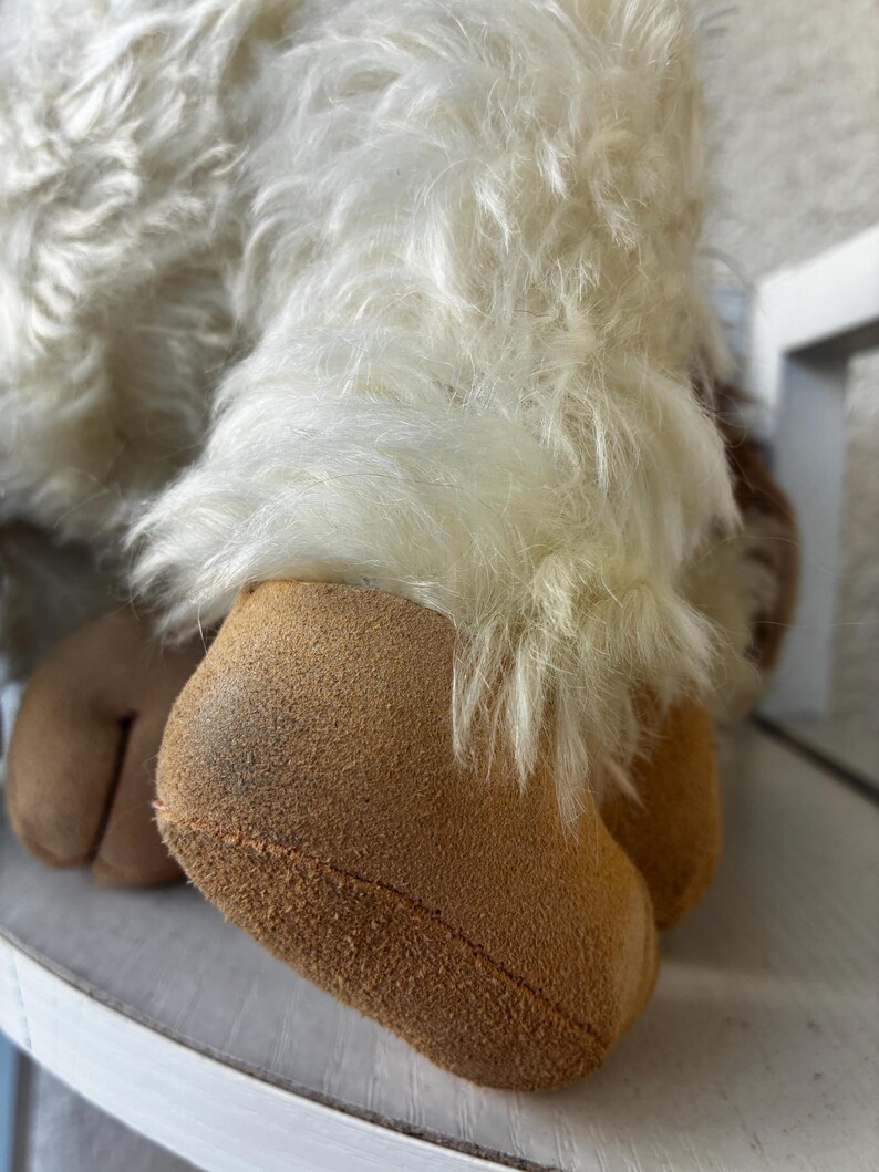May include: A plush toy featuring long, shaggy white faux fur covering the upper portion of the limbs, transitioning into smooth, tan-colored suede-like fabric at the hooves. The hooves are split, showing a distinct cleft design at the base.