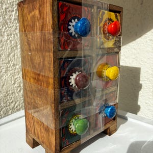 May include: A small wooden chest with six drawers, each featuring a unique ceramic knob in colors including blue, red, yellow, brown, and green. The drawer fronts are decorated with intricate patterns, and the entire unit is wrapped in clear plastic.