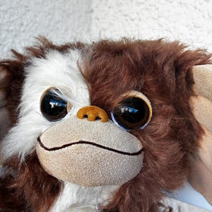 May include: A plush toy featuring a creature with long, shaggy brown and white fur. It has large, round, dark brown eyes, a small tan nose, and a wide, flat, light tan muzzle with a thin, dark horizontal line representing a closed mouth.