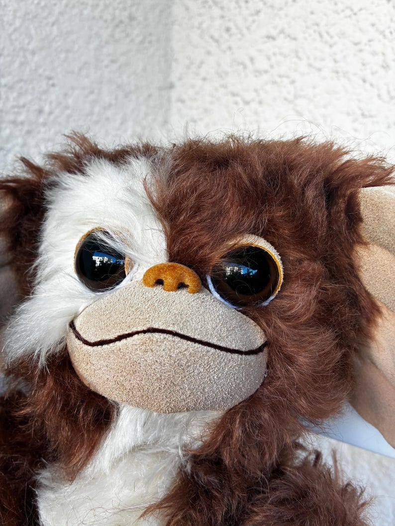 May include: A plush toy featuring a creature with long, shaggy brown and white fur. It has large, round, dark brown eyes, a small tan nose, and a wide, flat, light tan muzzle with a thin, dark horizontal line representing a closed mouth.