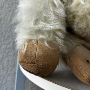 May include: A plush toy featuring long, shaggy off-white fur and tan suede-like fabric on the feet. The foot pad displays the partial black text 'cool' stitched onto the surface. The toy has a soft, textured appearance with visible seams.