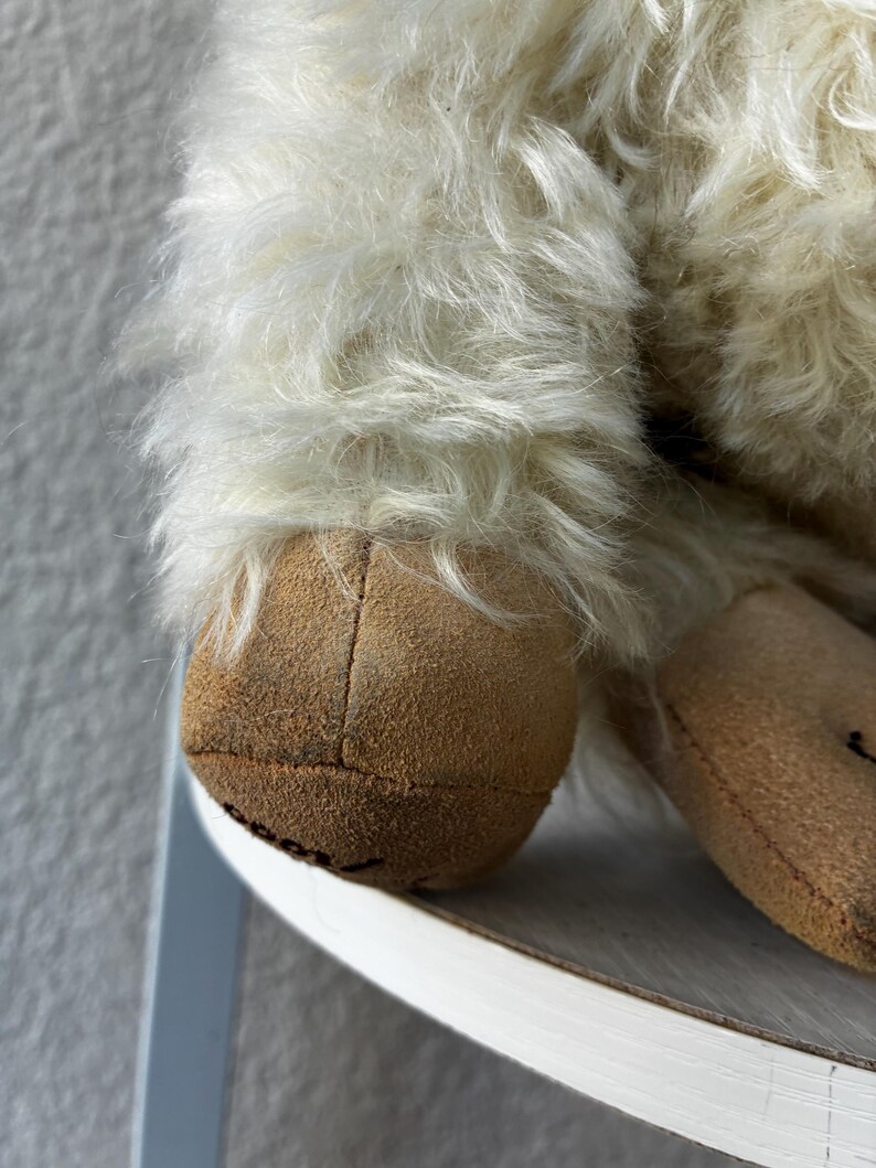 May include: A plush toy featuring long, shaggy off-white fur and tan suede-like fabric on the feet. The foot pad displays the partial black text 'cool' stitched onto the surface. The toy has a soft, textured appearance with visible seams.
