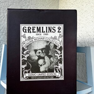 May include: A rectangular black box featuring a white label with the text GREMLINS 2 SINCE 1989 GENUINE MOHAIR COLLECTION GIZMO LIMITED EDITION. The label includes a black and white illustration of the character Gizmo surrounded by floral scrollwork.