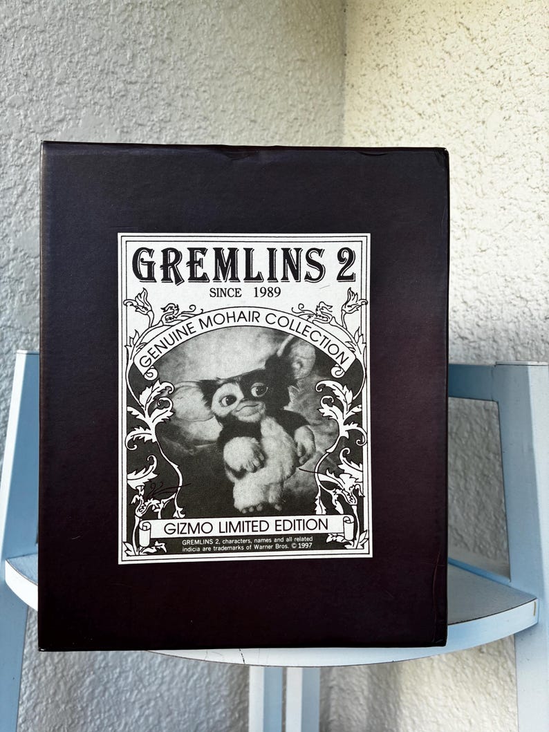 May include: A rectangular black box featuring a white label with the text GREMLINS 2 SINCE 1989 GENUINE MOHAIR COLLECTION GIZMO LIMITED EDITION. The label includes a black and white illustration of the character Gizmo surrounded by floral scrollwork.