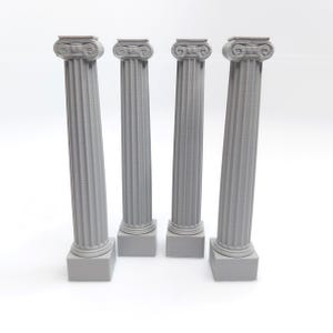 May include: Four miniature grey Ionic columns with detailed fluting and ornate capitals. The columns stand on square bases, creating a classical architectural theme. The columns are arranged in a row against a white background.