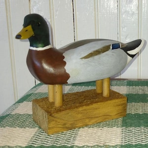 May include: Two wooden duck figurines. One duck has a brown body, green head, and yellow beak. The other duck is brown and tan. Both ducks are mounted on wooden bases and have wooden legs.