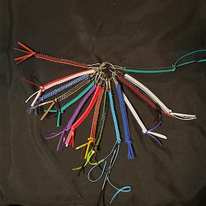 Może przedstawiać: Kolekcja 14 kolorowych, plecionych breloków do kluczy ze srebrnymi kółkami. Breloki są wykonane z kolorowych sznurków i mają około 15 cm długości.