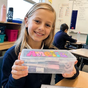 May include: A clear rectangular plastic pencil case with a hinged lid featuring the name Amelia in colorful, stylized bubble letters. The container is filled with various school supplies, including pens and markers, visible through the transparent material.