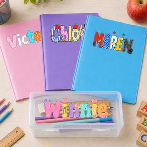 May include: Three notebooks in pink, purple, and blue with the names Victo, Chloe, and Maren printed on the covers. A clear plastic pencil case sits in front, labeled with the name Winnie, containing colored pencils and a wooden ruler.