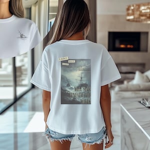 May include: White t-shirt with a small sketch of a sailboat on the left chest. The back features a graphic of a ship in a storm with the text "WILL SINK BENEATH THE WAVES". The shirt is paired with denim shorts.