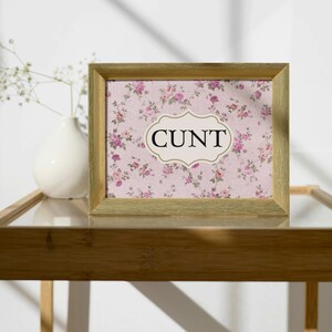 May include: A gold framed picture with a pink floral pattern and the word "CUNT" in a white oval.