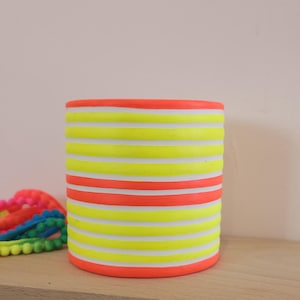 Puede incluir: Un jarrón cilíndrico de cerámica con rayas horizontales amarillas neón, blancas y naranjas. El jarrón está sobre una superficie de madera, con una guirnalda de pompones coloridos parcialmente visible. Es un objeto decorativo.