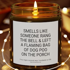 May include: A brown glass candle jar with a white label that reads "Smells Like Someone Rang The Bell & Left A Flaming Bag Of Dog Poo On The Porch". The candle is 9 oz. and made with vegan coconut wax. The label also includes the website "SmellsLikeVibes.com".