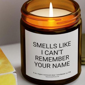 Peut inclure: Un bocal en verre brun contenant une bougie avec une étiquette blanche sur laquelle est inscrit "Smells Like I Can't Remember Your Name". La bougie est allumée et le bocal est posé sur une surface jaune. L'étiquette indique également "9 oz. vegan coconut wax | handmade in USA" et "SmellaLikeVibes.com".