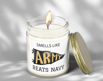 Può includere: Una candela accesa in un barattolo di vetro trasparente con coperchio dorato. L'etichetta recita "Smells Like Army Beats Navy" in lettere nere e dorate. La candela è su una superficie bianca.