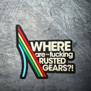 May include: A black sticker with the text "WHERE are the fucking RUSTED GEARS?!" in white. A colorful design of red, yellow, green, and blue lines is on the left side of the sticker.