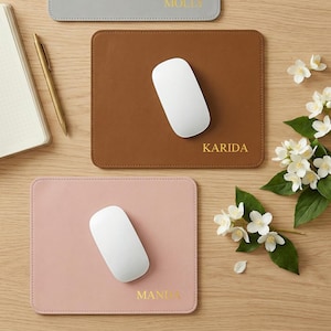 May include: Three rectangular mouse mats in grey, brown, and pink, each with a white mouse. The grey mat has the name "MOLLY" in gold, the brown mat has "KARIDA", and the pink mat has "MANIA". A notebook, pen, and flowers are also present.