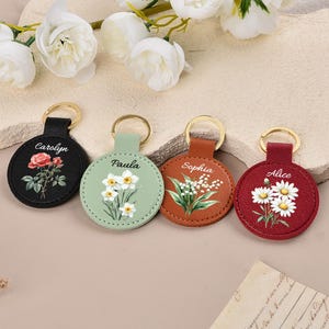 Pode incluir: Quatro chaveiros redondos de couro em preto, verde, castanho e vermelho, cada um com um anel dourado. Os chaveiros apresentam desenhos florais e nomes: Carolyn, Paula, Sophia e Alice. Flores brancas e vegetação são pintadas nos chaveiros.