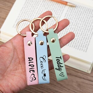 Puede incluir: Tres llaveros de cuero personalizados con anillos dorados. Los llaveros son rosa, azul claro y verde claro. Los nombres "Alfie", "Coco" y "Teddy" están escritos en los llaveros.