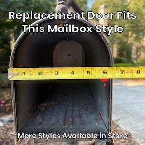 May include: A dark metal mailbox with an open front, featuring a yellow Stanley measuring tape across the opening showing a width of approximately 8 inches. Text reads: Replacement Door Fits This Mailbox Style and More Styles Available in Store.