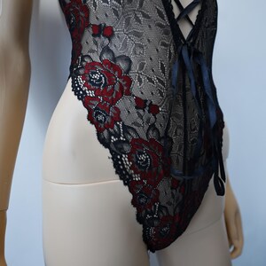 Puede incluir: Body de encaje negro con bordado floral rojo. El body tiene un escote pronunciado y un detalle de encaje en la parte delantera.