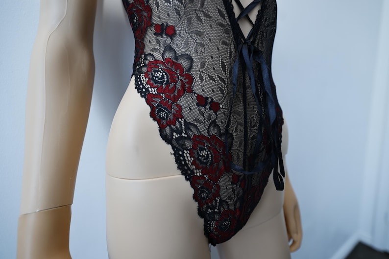 Puede incluir: Body de encaje negro con bordado floral rojo. El body tiene un escote pronunciado y un detalle de encaje en la parte delantera.