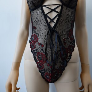Puede incluir: Body de encaje negro con bordado floral rojo y un frente con cordones.