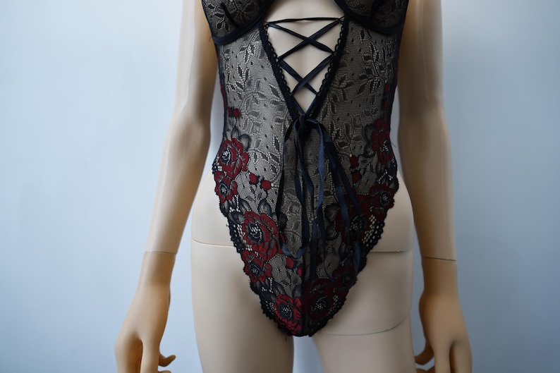 Puede incluir: Body de encaje negro con bordado floral rojo y un frente con cordones.