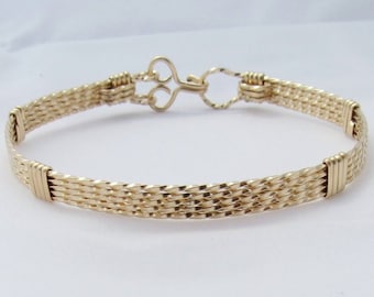 Könnte beinhalten: Ein geflochtenes Armband aus goldfarbenem Draht mit einem schlüsselförmigen Verschluss.